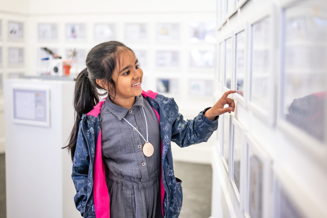 A young girl with dark hair in a ponytail smiles whilst pointing at framed pictures on a white gallery wall. She wears a grey dress and a blue jacket with pink lining.