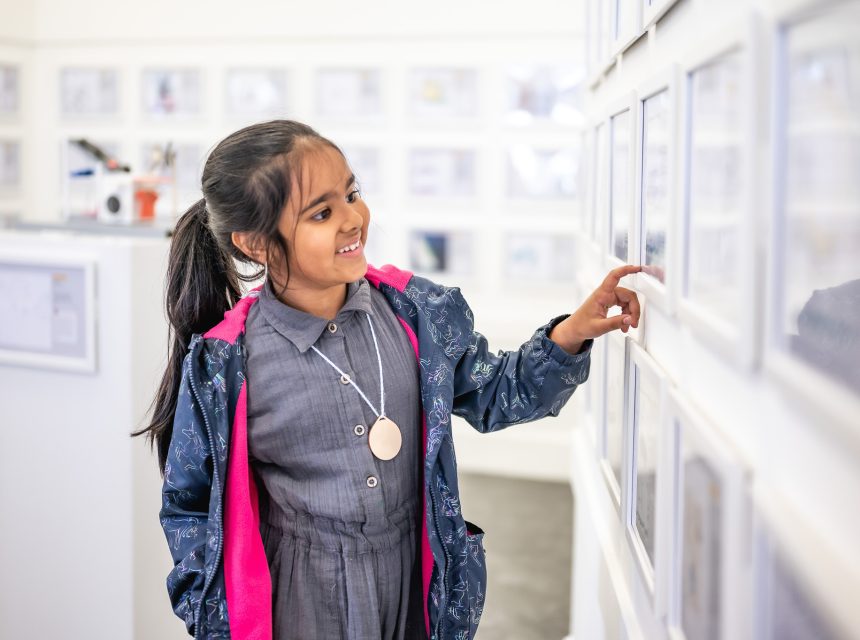 A young girl with dark hair in a ponytail smiles whilst pointing at framed pictures on a white gallery wall. She wears a grey dress and a blue jacket with pink lining.