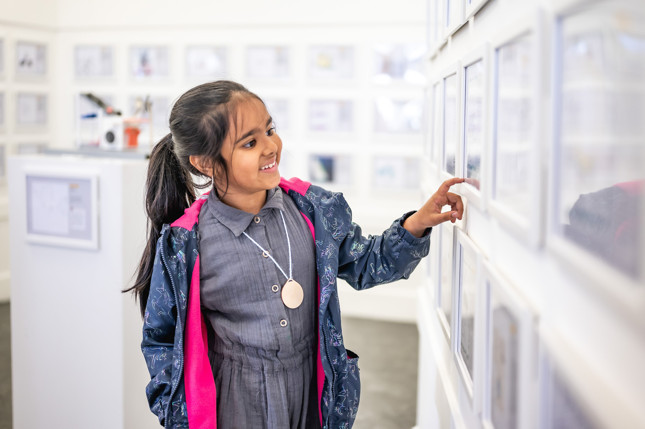 A young girl with dark hair in a ponytail smiles whilst pointing at framed pictures on a white gallery wall. She wears a grey dress and a blue jacket with pink lining.
