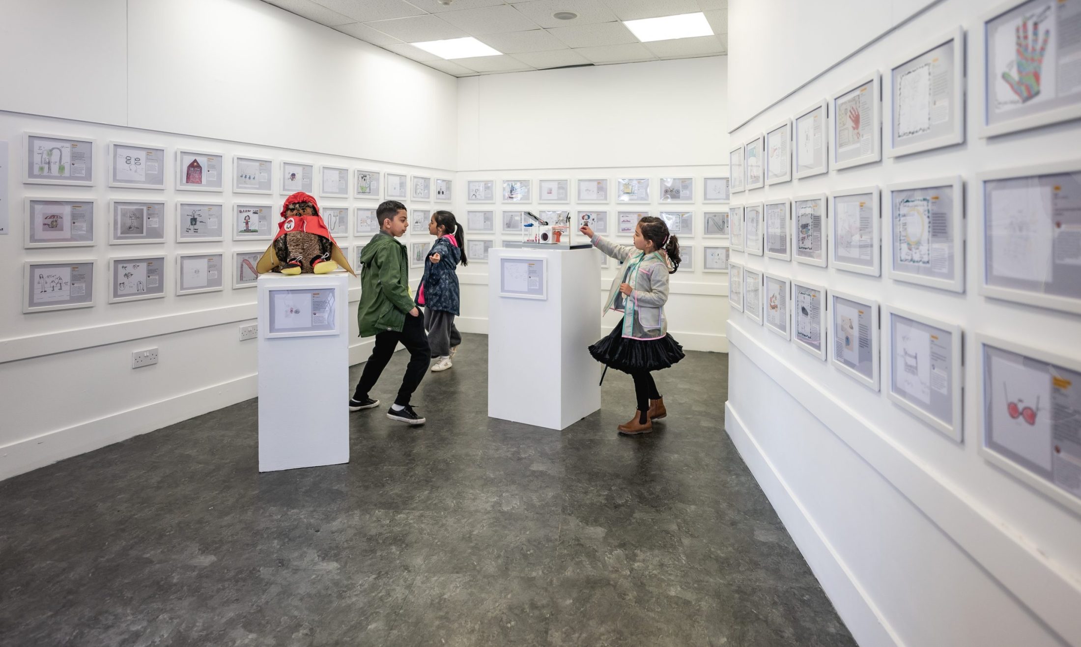 Four children look at framed artwork displayed on the white walls of a brightly lit art gallery. Some children interact with art on plinths at the centre of the room. The atmosphere appears lively and curious.