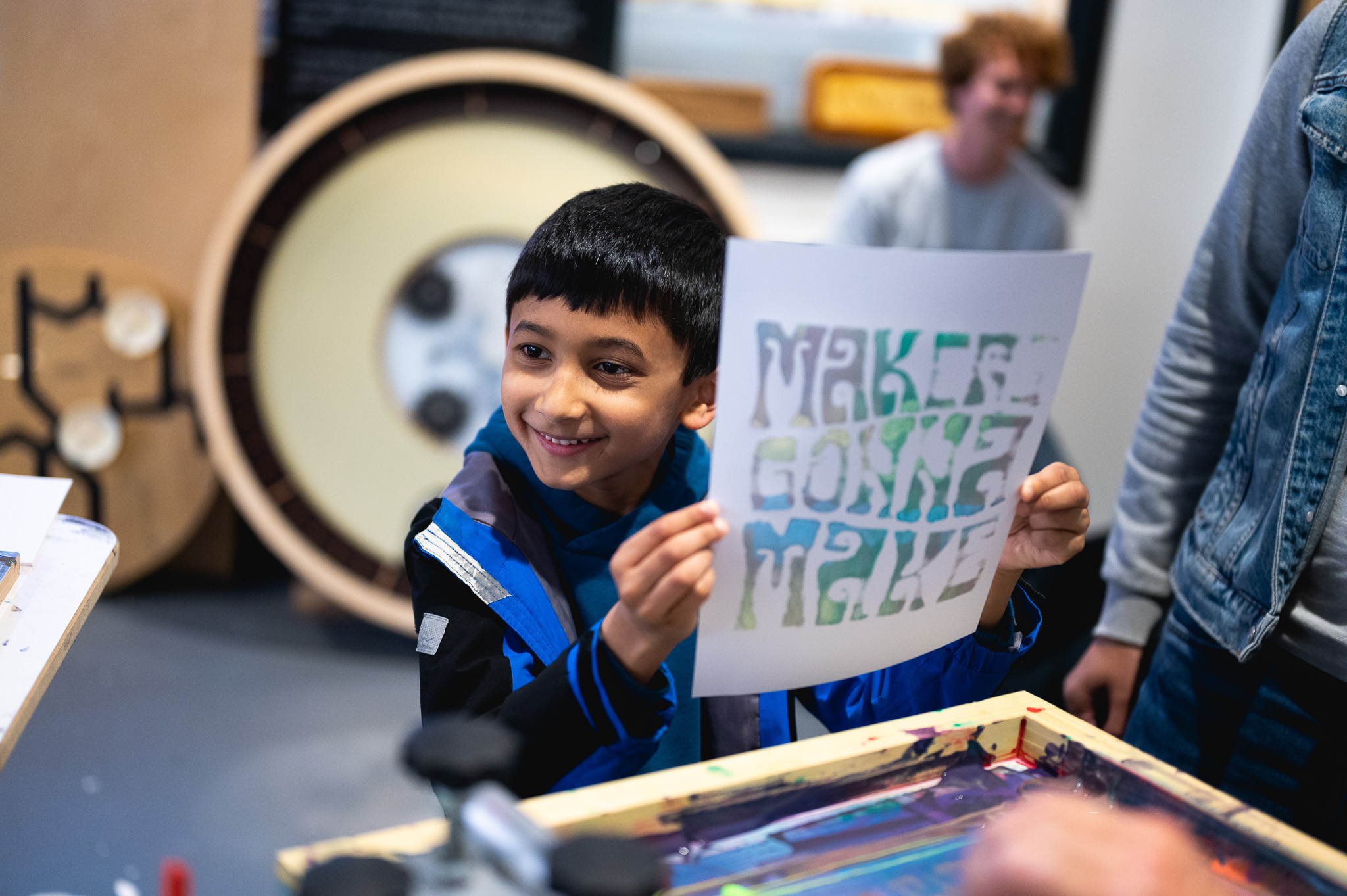 A smiling boy holds up a paper that says MAKE & GONNA MAKE in colourful letters, showing his artwork at a creative workshop. People and art materials are visible in the background.