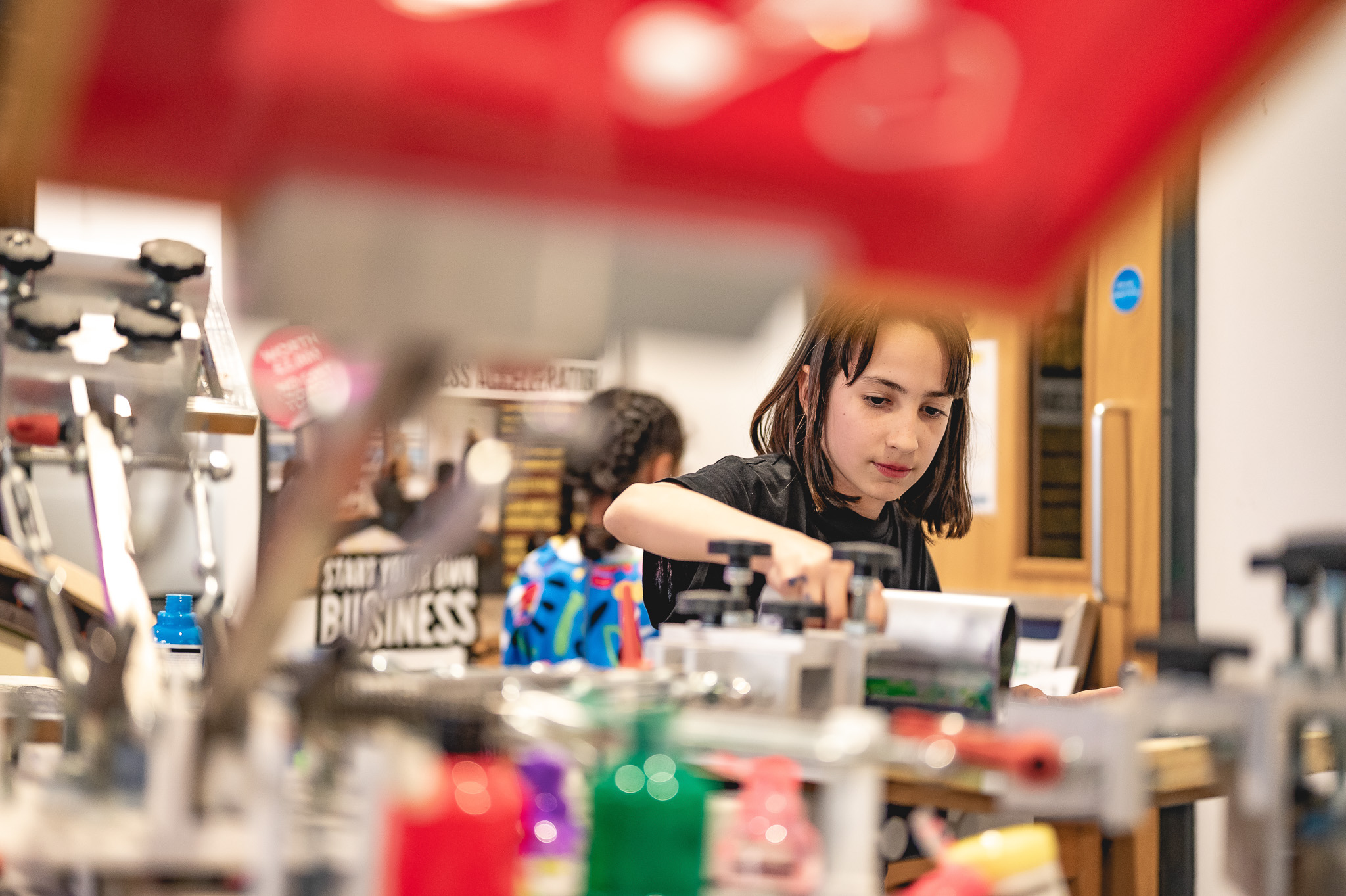 A child focuses intently while working with colourful equipment and tools in a busy, creative workshop or classroom setting.