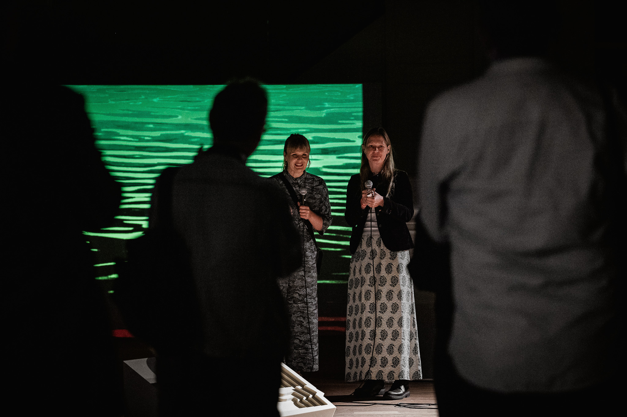 Two women stand and speak in front of a green-lit screen displaying water ripples, whilst silhouetted people watch them in a dimly lit room.