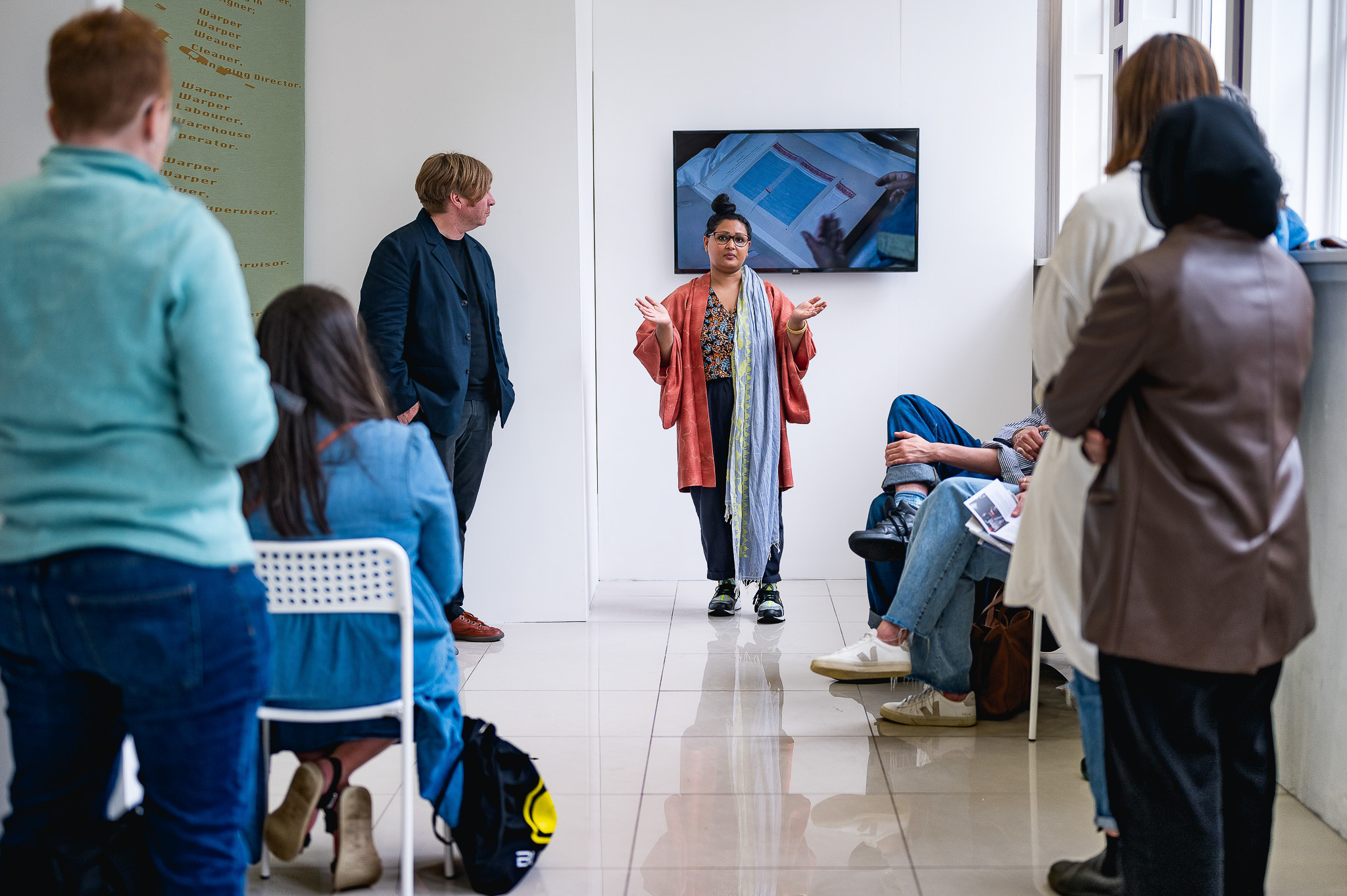 A person stands in front of a group, gesturing whilst speaking. Another person stands nearby. The audience is seated and facing them. A screen displaying images is on the wall behind the speakers. The setting appears to be a modern room.