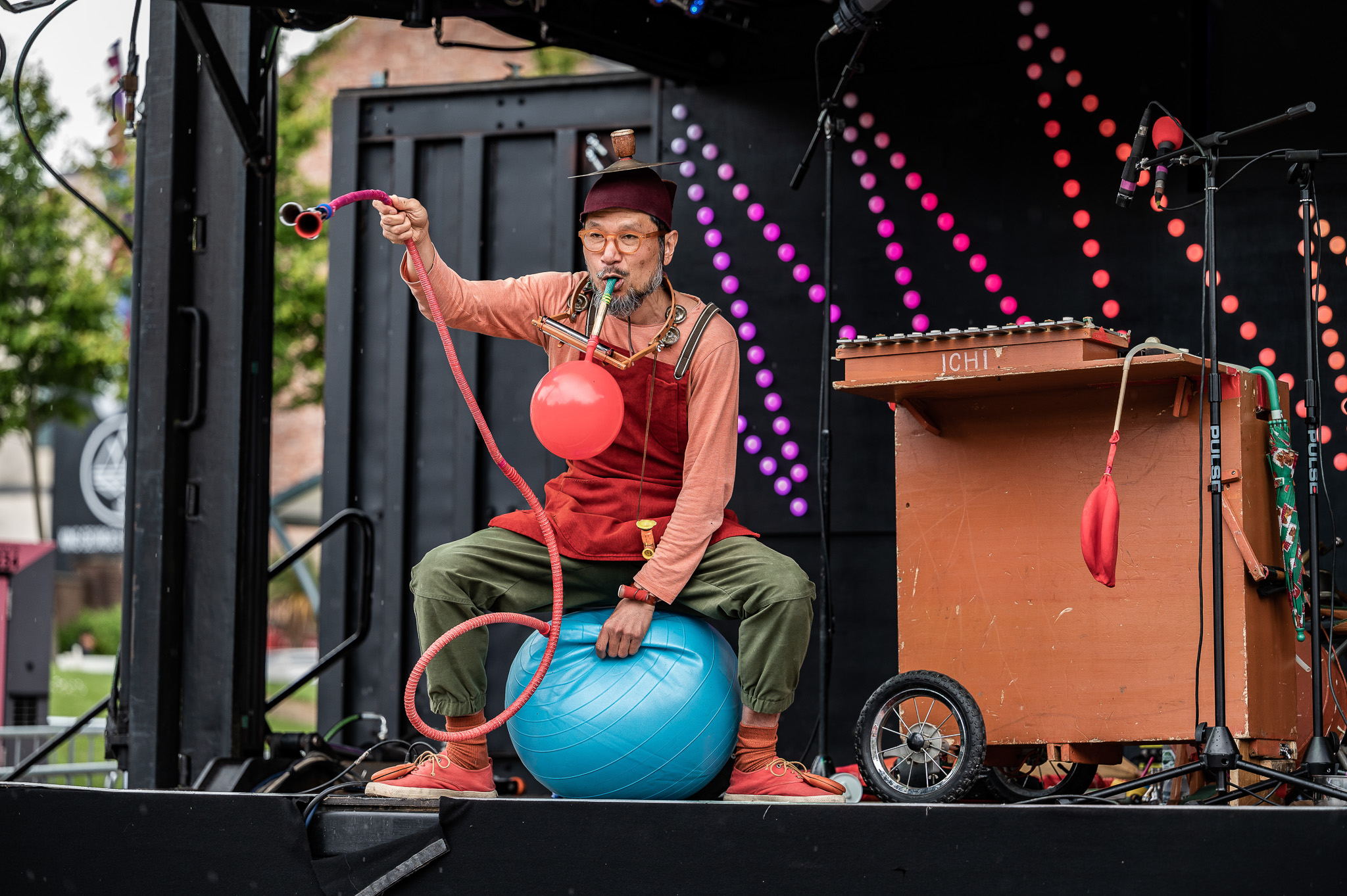 A performer in colourful clothes sits on a blue exercise ball, inflating a red balloon with a pump. The performer is on an outdoor stage with a wooden cart and colourful lights in the background.