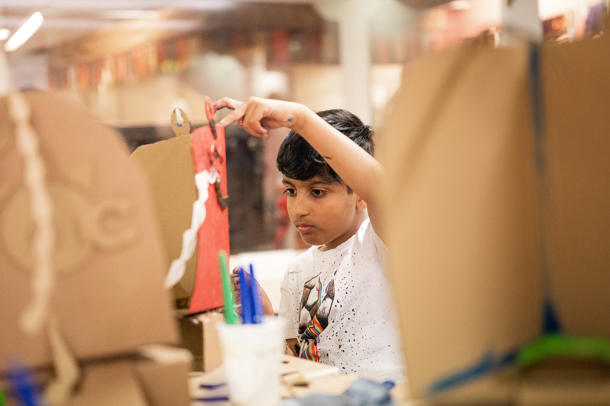 A young boy focuses intently whilst decorating a cardboard structure with paint and other craft materials in a creative classroom setting.