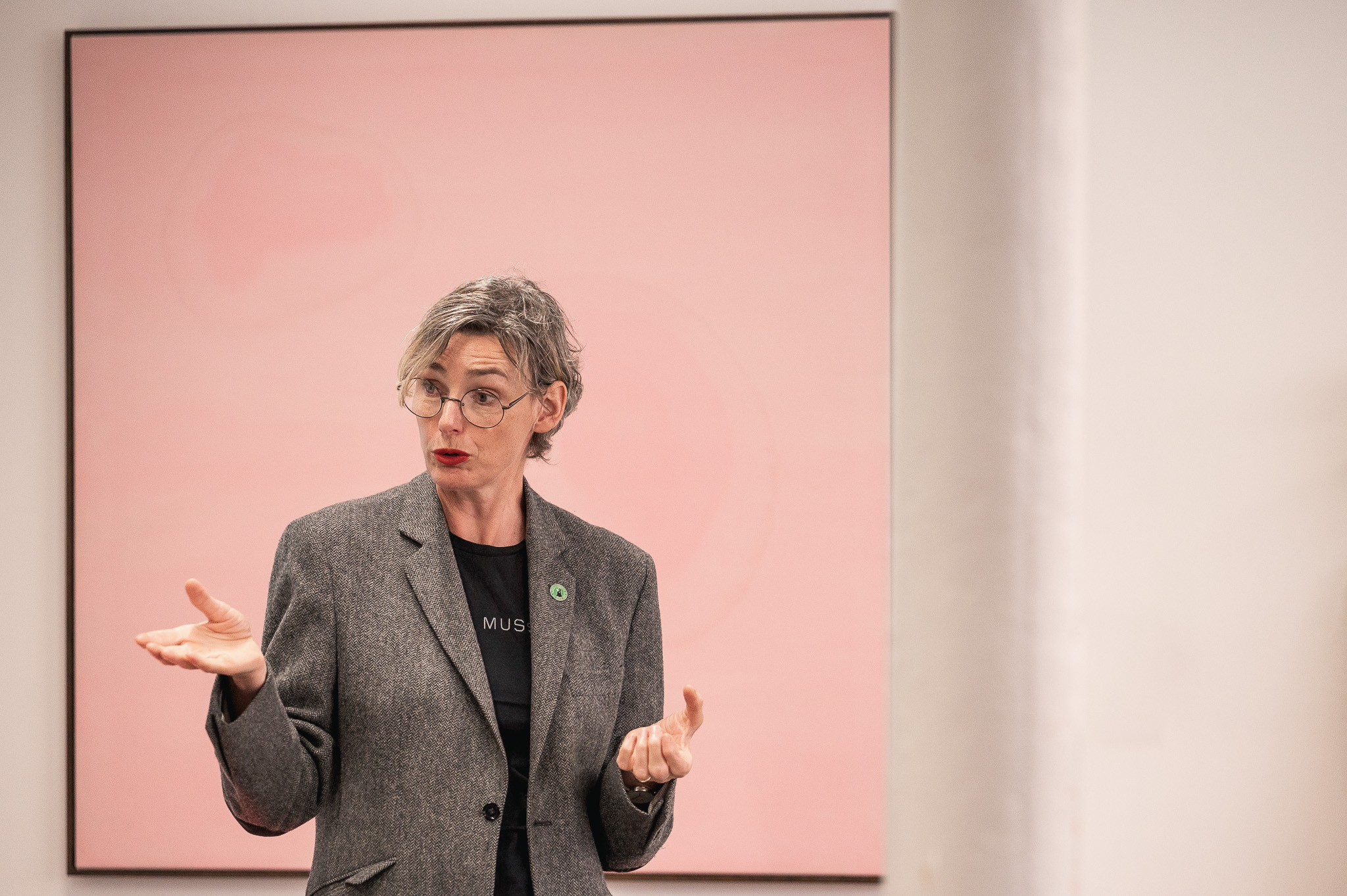 Jacqueline Donachie speaks passionately while gesturing with open hands. A large pink abstract painting hangs behind them.