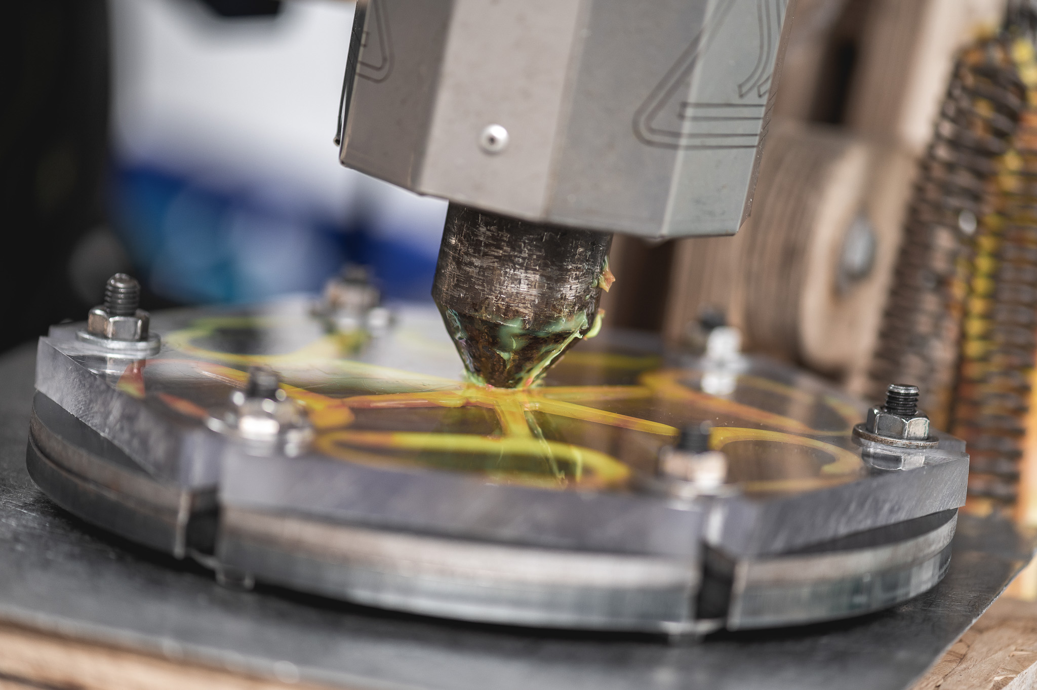 A close-up of a 3D printer nozzle extruding yellow filament onto a circular platform, creating a layered pattern. Bolts secure the platform, and parts of the 3D printer are visible in the background.