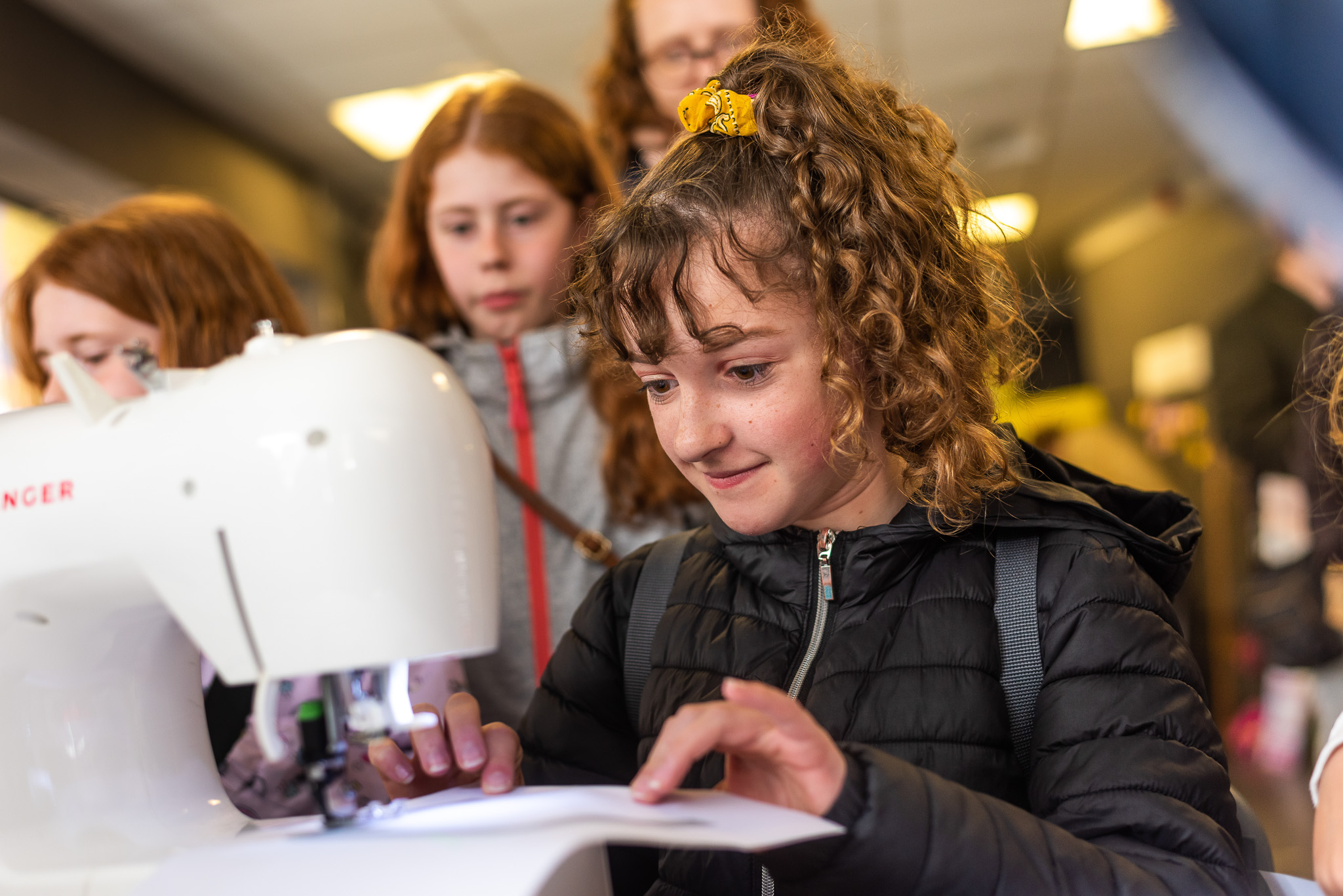 A girl with curly hair and a yellow scrunchie focuses on using a sewing machine whilst other children watch in the background. She wears a black jacket and smiles slightly as she guides fabric under the needle.