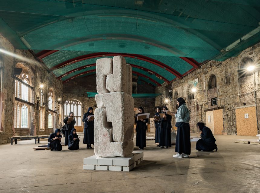 A group of people dressed in black observe and sketch a tall stone sculpture inside a spacious, rustic hall with arched windows, exposed brick walls, and a high, turquoise and red ceiling.