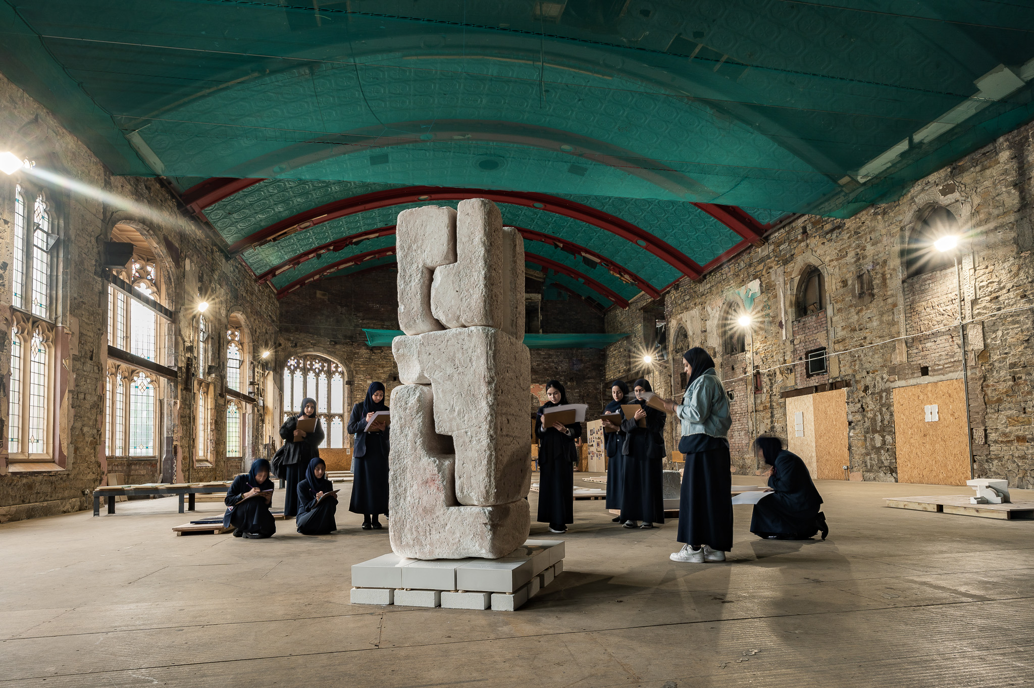 A group of people dressed in black observe and sketch a tall stone sculpture inside a spacious, rustic hall with arched windows, exposed brick walls, and a high, turquoise and red ceiling.