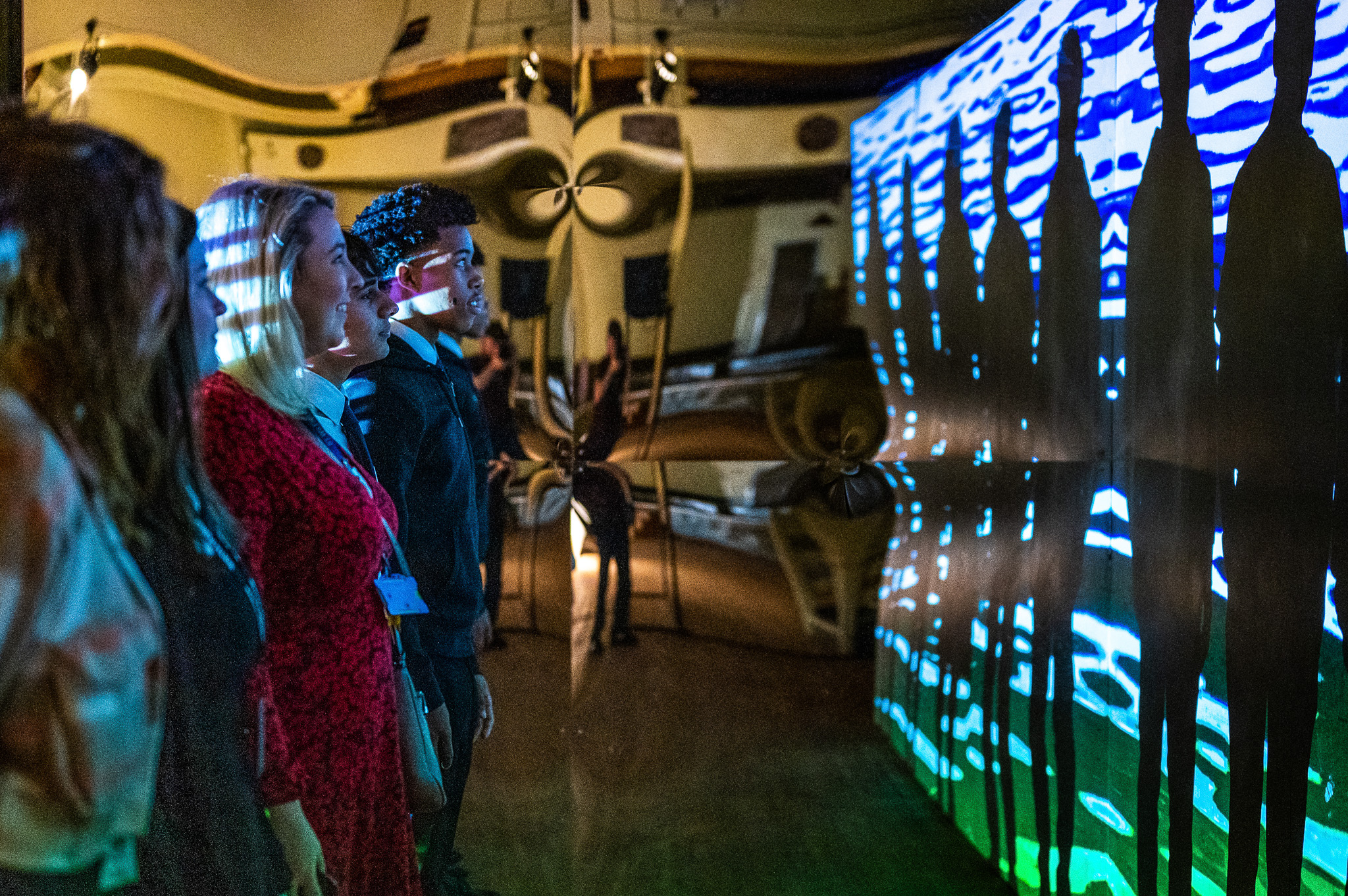 Several people stand facing a brightly lit, interactive wall display with colourful patterns and silhouettes, in a dimly lit room with reflective, distorted mirrors.