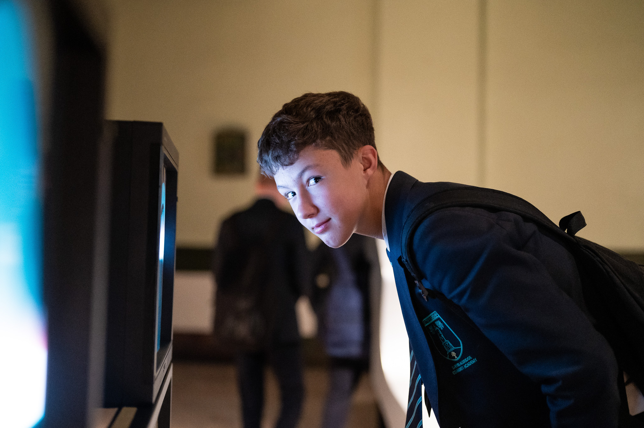A teenage boy in a school uniform with a rucksack leans forward to look closely at a lit display screen, smiling slightly. The background is softly blurred with people walking past.