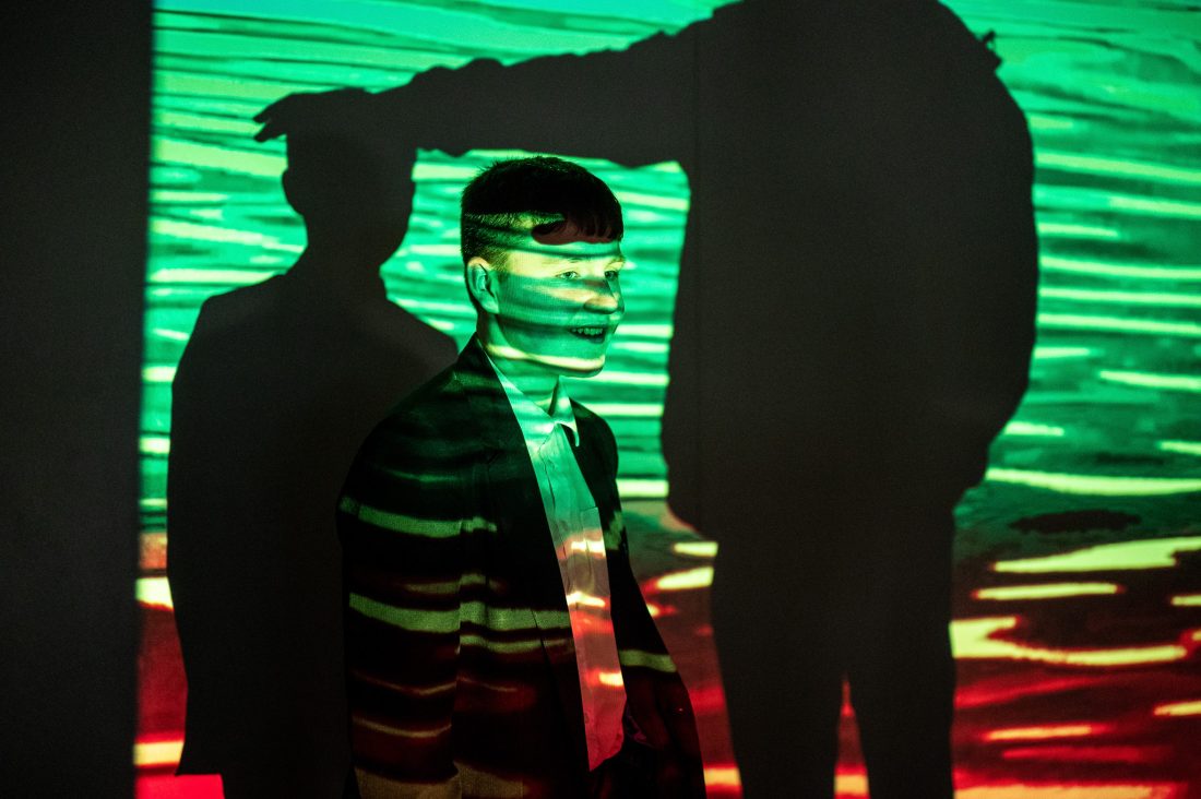 A person stands against a wall with green and red light patterns projected onto them. Large shadows of another figure appear beside and over the person, creating a dramatic, surreal effect.