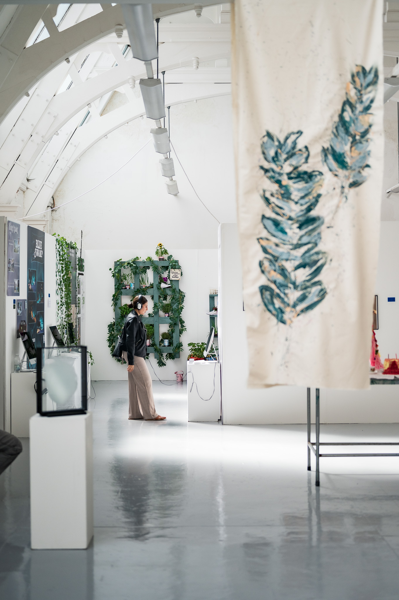 A person walks through a bright, modern art gallery with white walls, leafy green plants, and a large hanging fabric with blue-green leaf prints in the foreground.