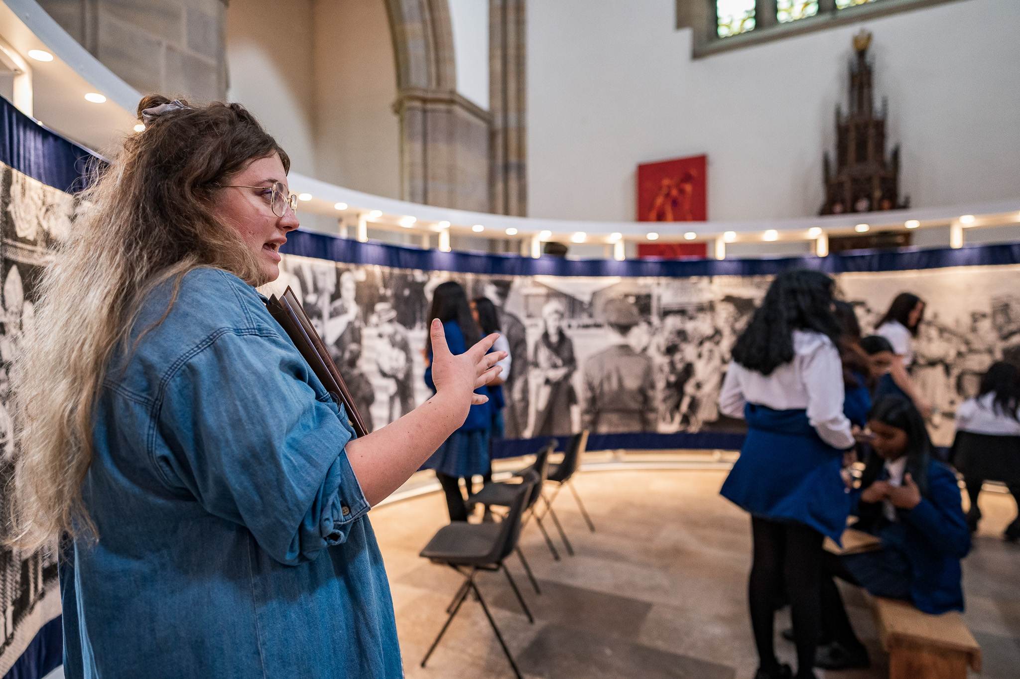 A woman speaking gestures with her hand in a church-like space, whilst a group of girls in blue uniforms gather and talk near a large, curved, black-and-white photo display in the background.