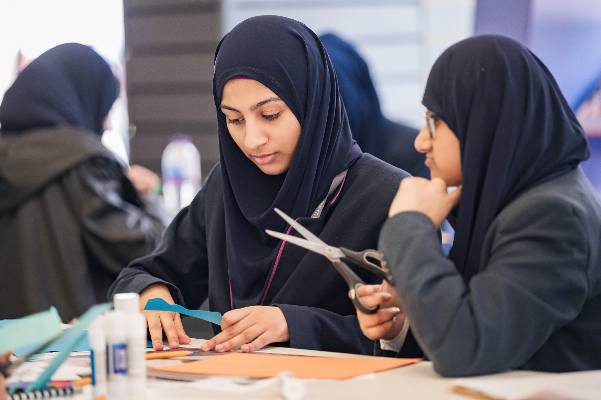 Two girls wearing hijabs sit at a table working on an arts and crafts project with paper, scissors, and glue, focused and collaborating in a classroom setting.