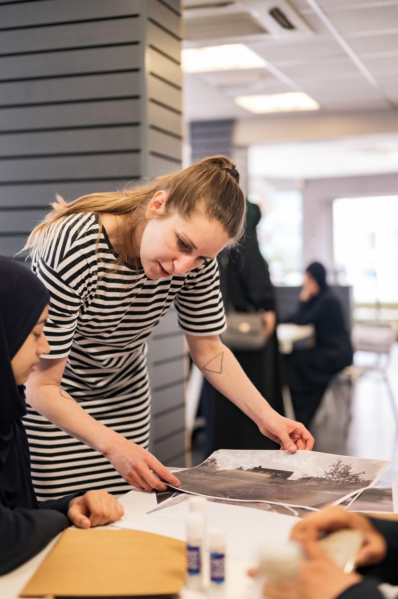 A woman in a striped dress leans over a table, showing a large black-and-white photo to two people seated. Others are visible in the background in a bright, modern room.