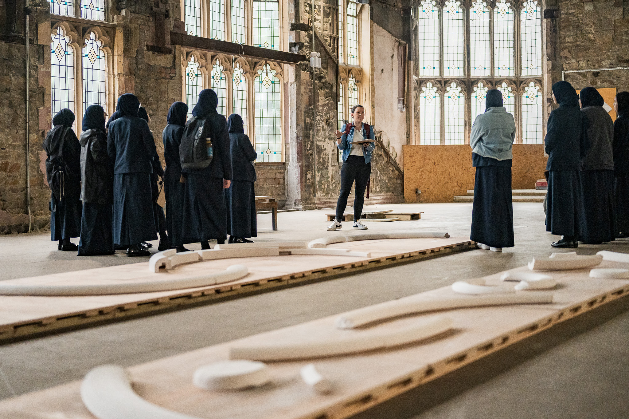 A group of students in dark clothing stand in a large, historic hall with stained glass windows, listening to a person speaking. Sculptural art pieces lie on wooden panels on the floor.