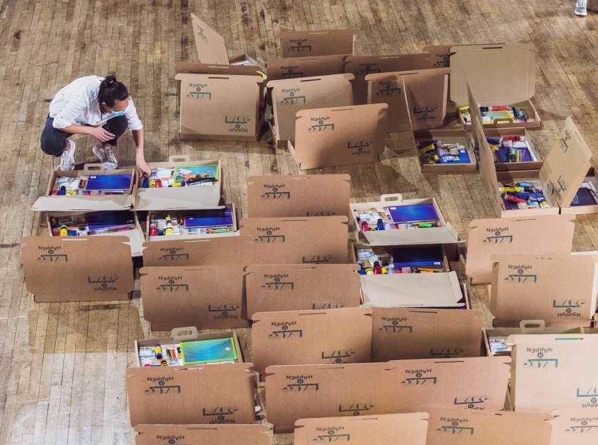 A person arranges items in large open cardboard boxes on a wooden floor; the boxes contain various art supplies.