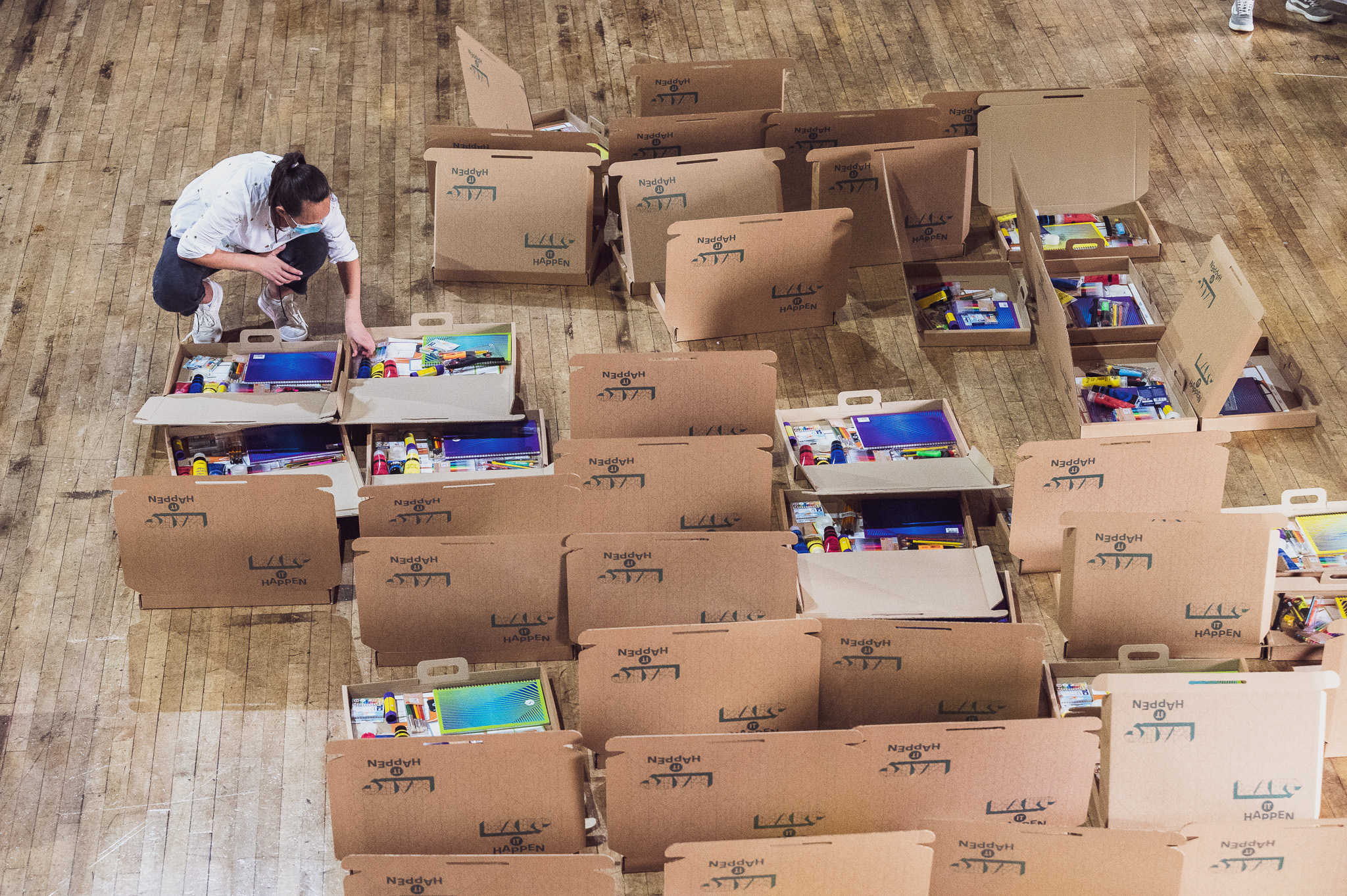 A person arranges items in large open cardboard boxes on a wooden floor; the boxes contain various art supplies.