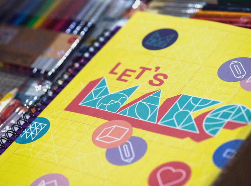 A yellow notebook with LET'S MAKE in bold, colourful geometric letters on the cover, surrounded by scattered art supplies like coloured pens and markers. Various geometric shapes decorate the notebook.
