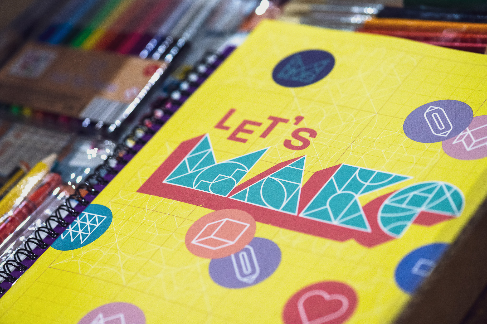 A yellow notebook with LET'S MAKE in bold, colourful geometric letters on the cover, surrounded by scattered art supplies like coloured pens and markers. Various geometric shapes decorate the notebook.