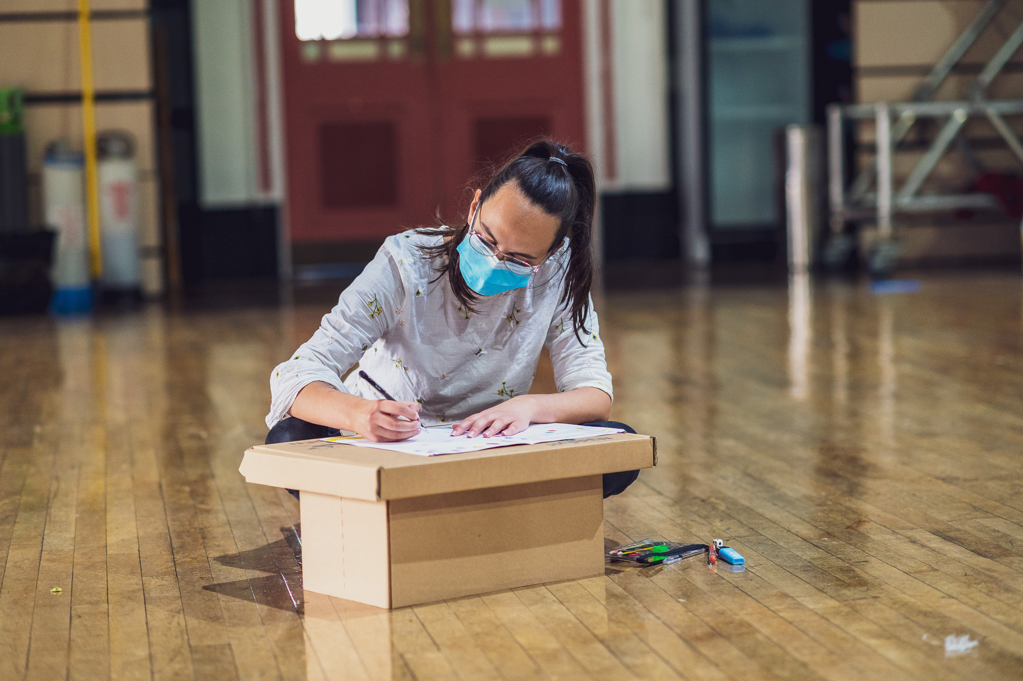 A young person wearing a blue face mask sits on the floor in a spacious room, drawing on paper placed on a cardboard box, with coloured pens scattered nearby.
