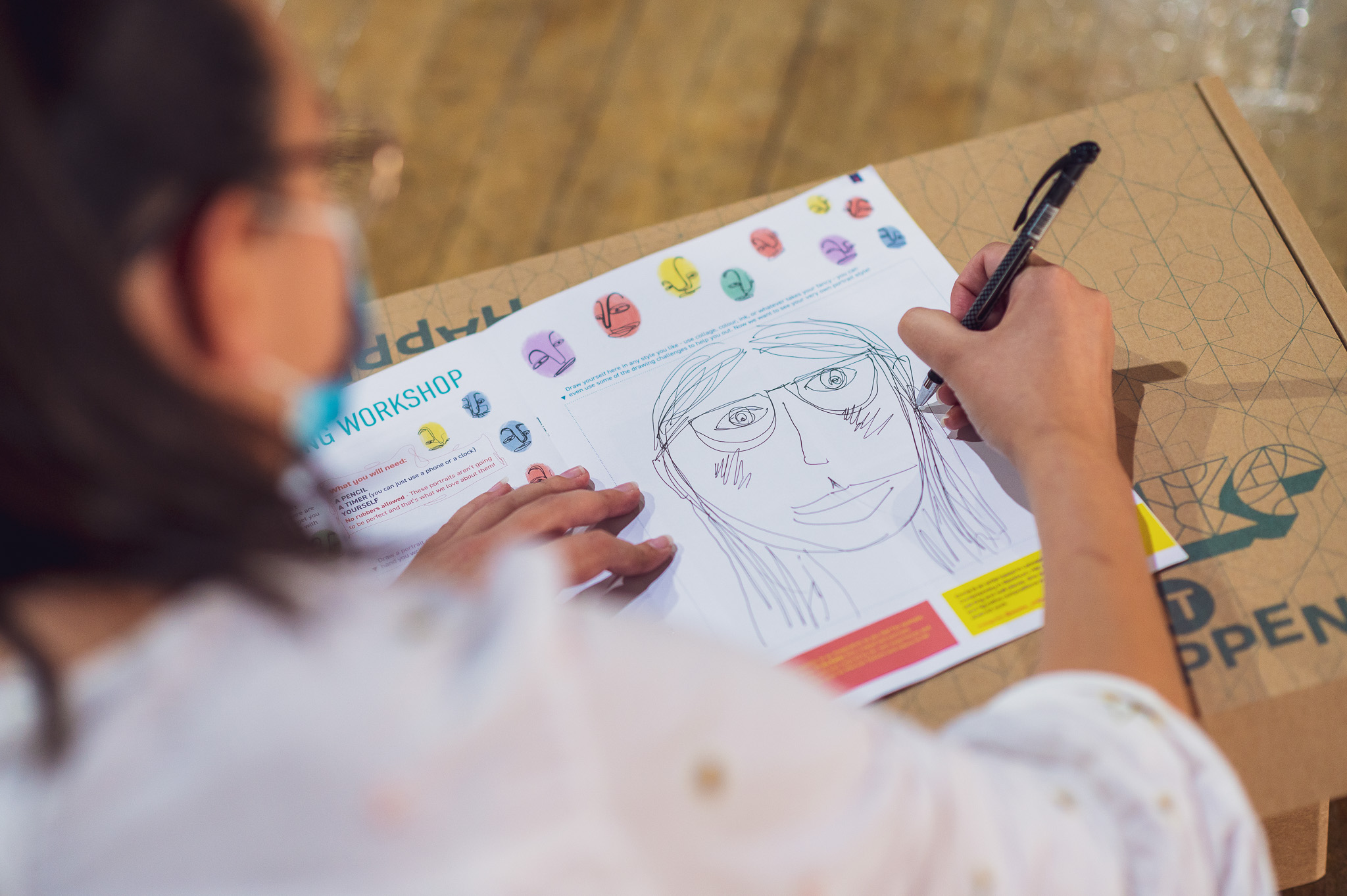 A person wearing glasses and a mask draws a large face on an activity sheet, with colourful illustrated faces and text visible on the page, sitting at a cardboard box table.