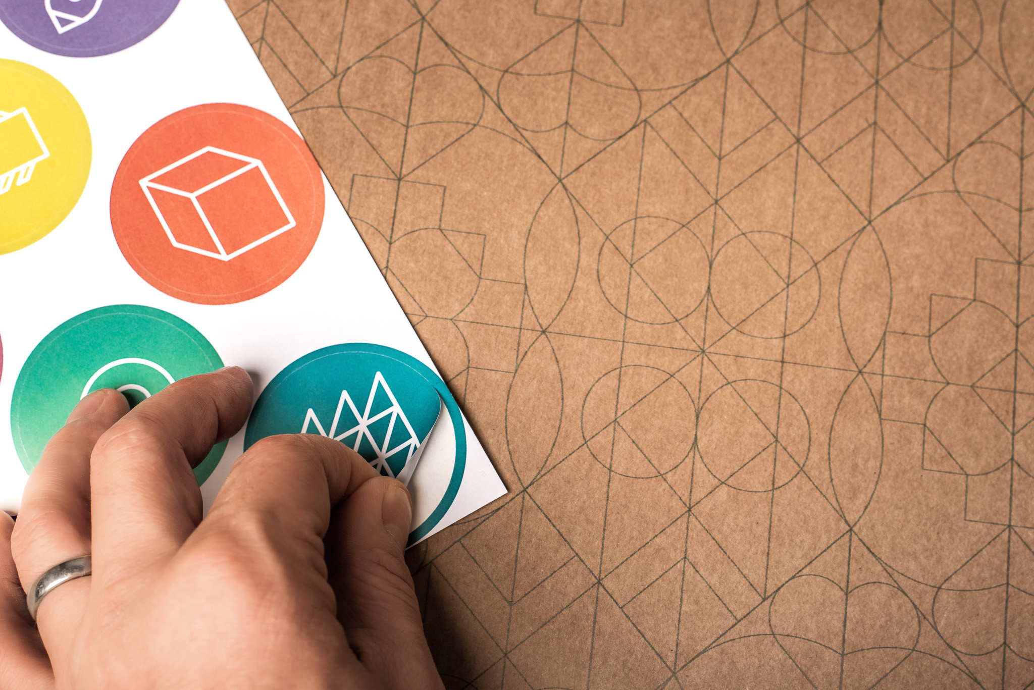 A hand with a silver ring peels a blue geometric sticker from a sheet of colourful round stickers, all featuring white line art icons, over a patterned brown surface.