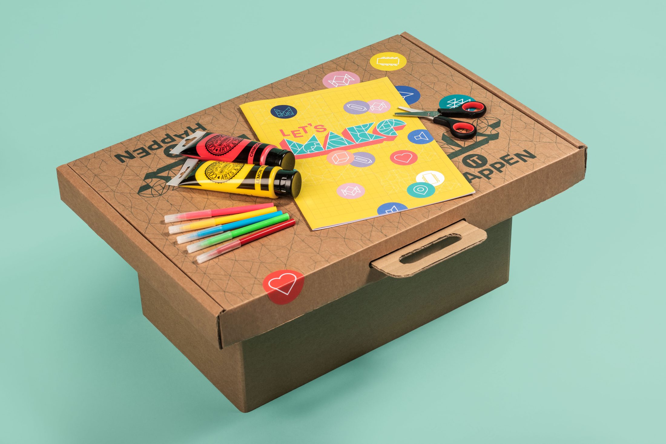 A cardboard box serves as a table for art supplies, including coloured markers, a glue stick, scissors, a yellow notebook, and stickers, all set on a light teal background.