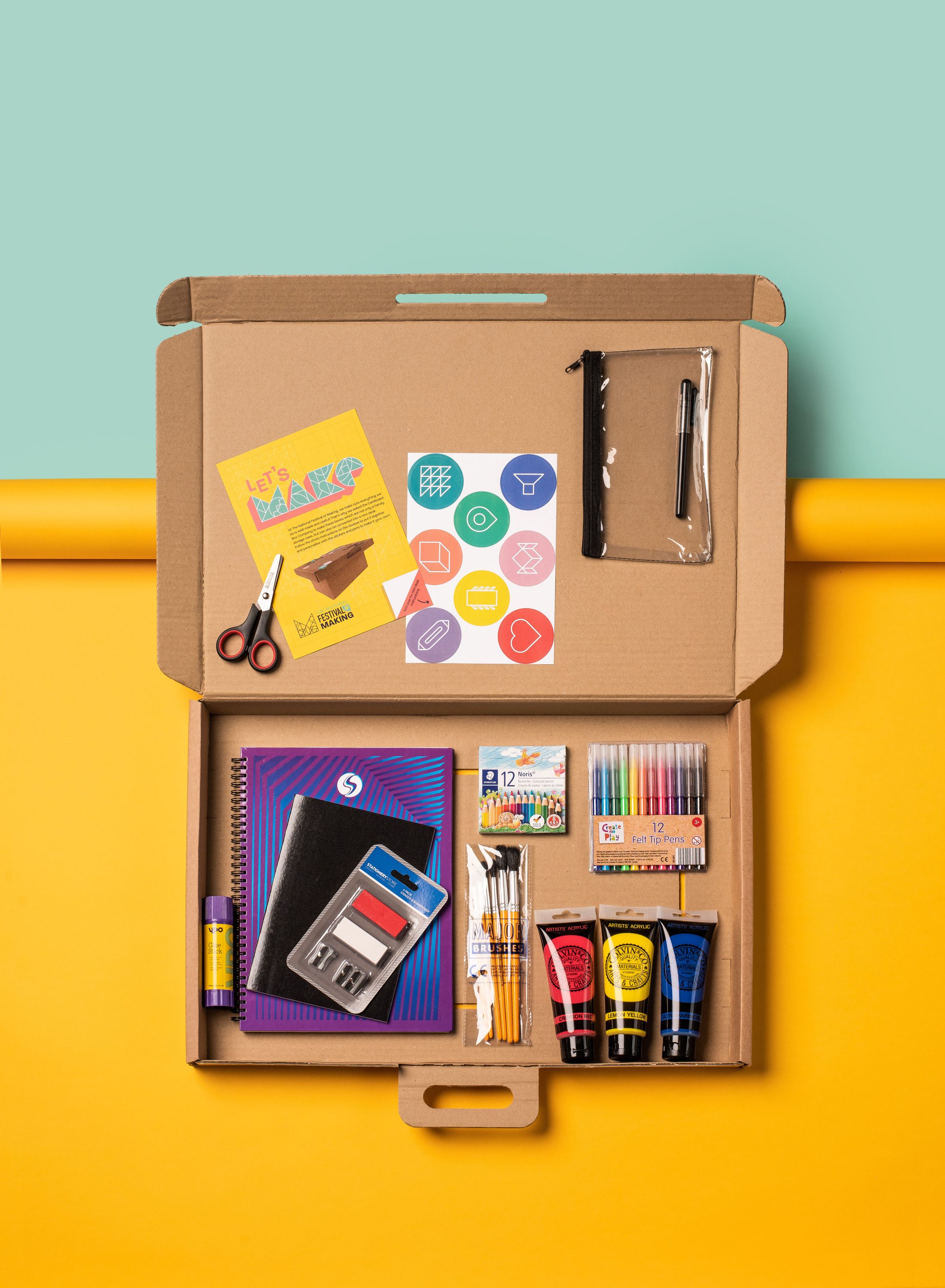 Open cardboard box on a yellow surface containing art supplies: scissors, notebook, felt-tip pens, paints, paintbrushes, glue stick, rubber, sharpener, and colourful leaflets with icons. Background is split yellow and mint green.