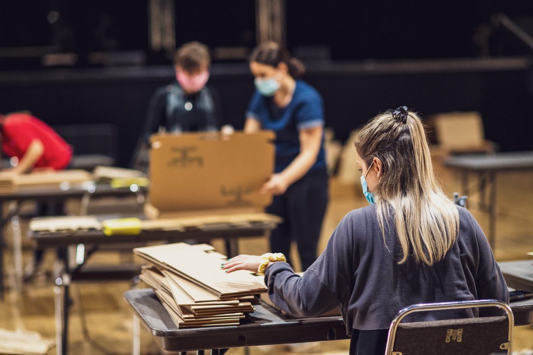 Four people wearing masks are assembling cardboard boxes at tables in a spacious indoor setting. The focus is on a woman with long hair in the foreground, whilst others work in the background.