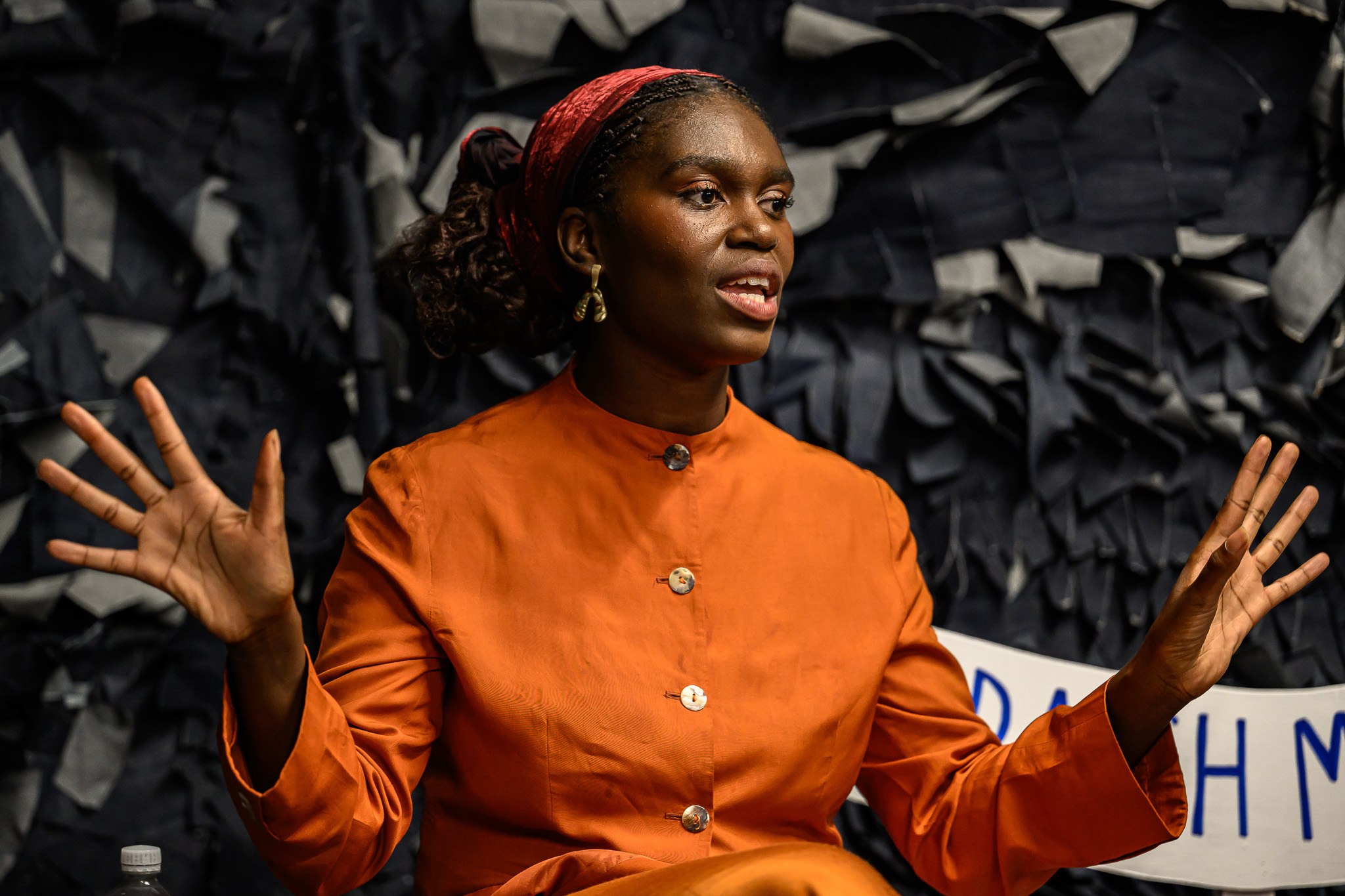 A woman in an orange dress with a red headband gestures with both hands whilst speaking, seated in front of a dark, textured backdrop.