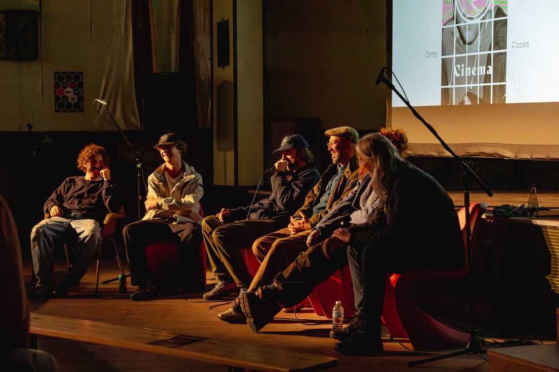 Six people sit on stage in a dimly lit room, participating in a panel discussion. Microphones are set up and a projected screen behind them displays the word Cinema.