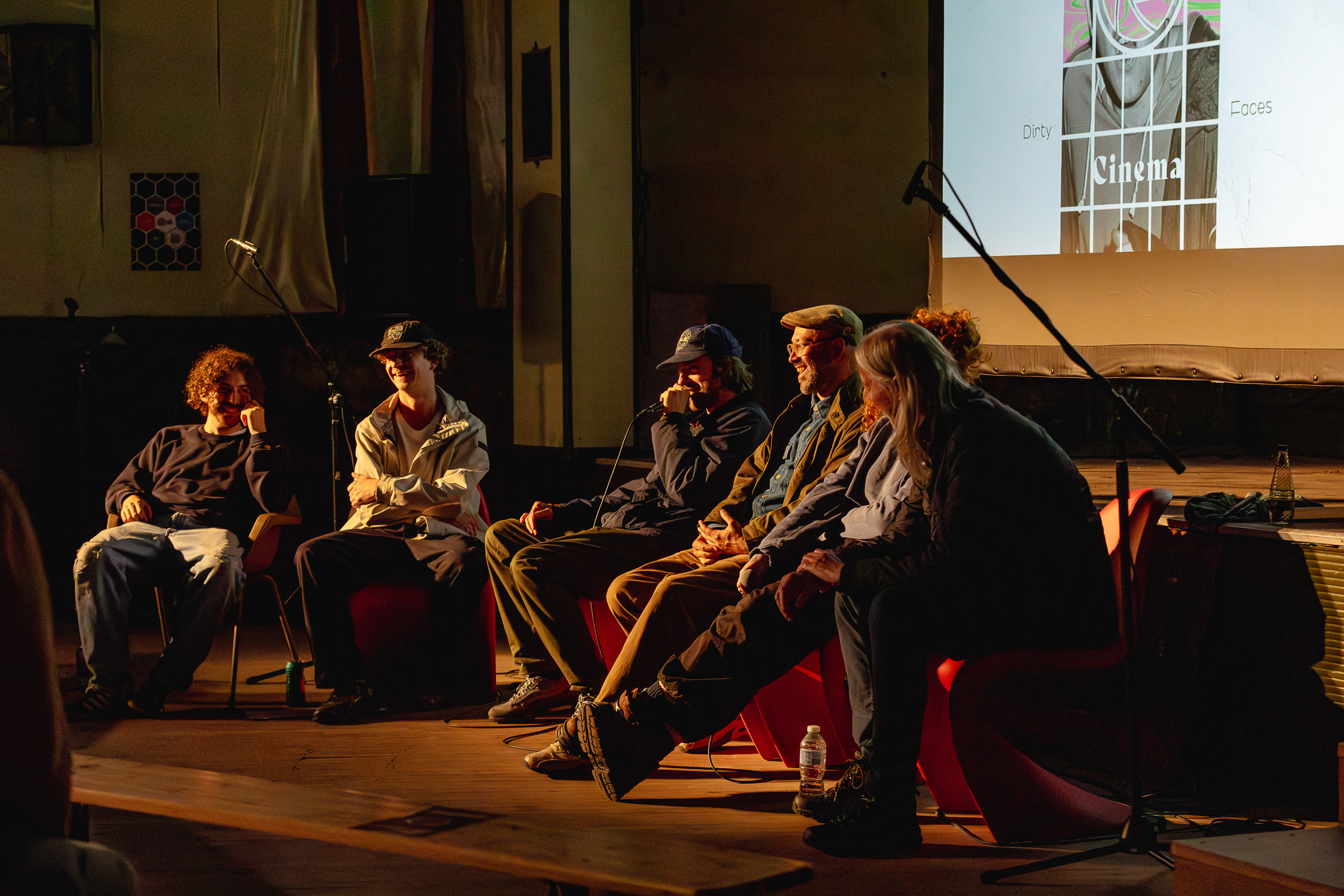 Six people sit on stage in a dimly lit room, participating in a panel discussion. Microphones are set up and a projected screen behind them displays the word Cinema.