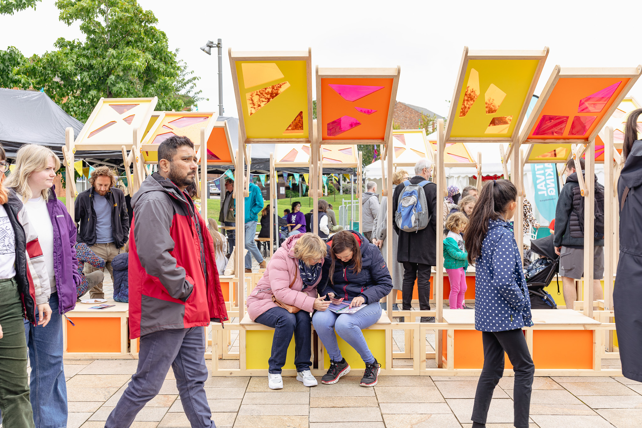 People gather outdoors at a public event; some sit under colourful, geometric canopies while others walk by. Tents and trees are visible in the background, creating a vibrant, lively atmosphere.