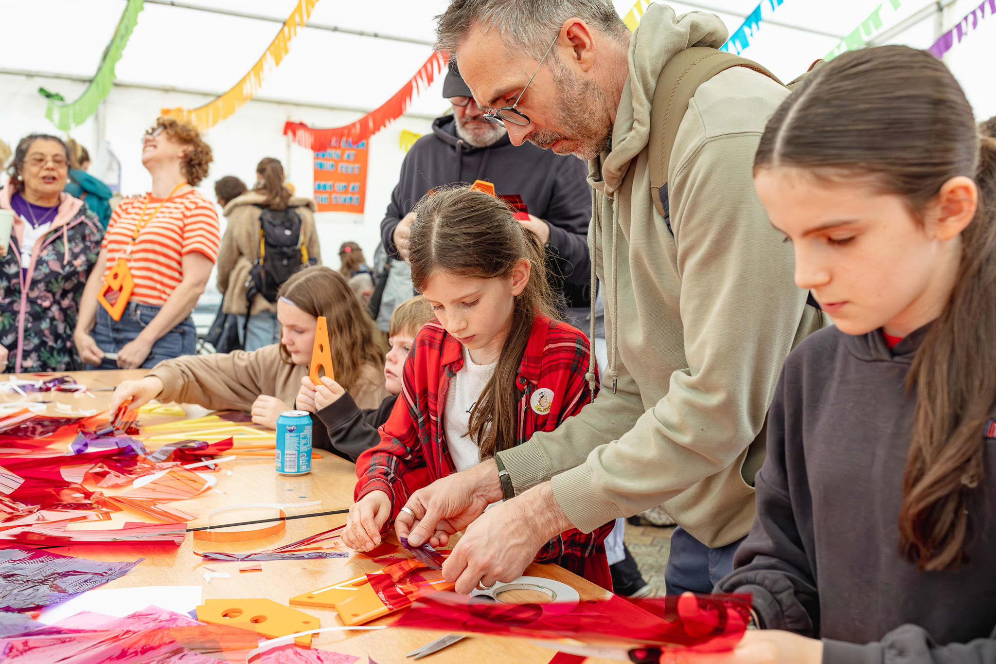A man assists children with a colourful craft project on a crowded table, surrounded by vibrant paper and festive decorations under a lively, cheerful atmosphere.