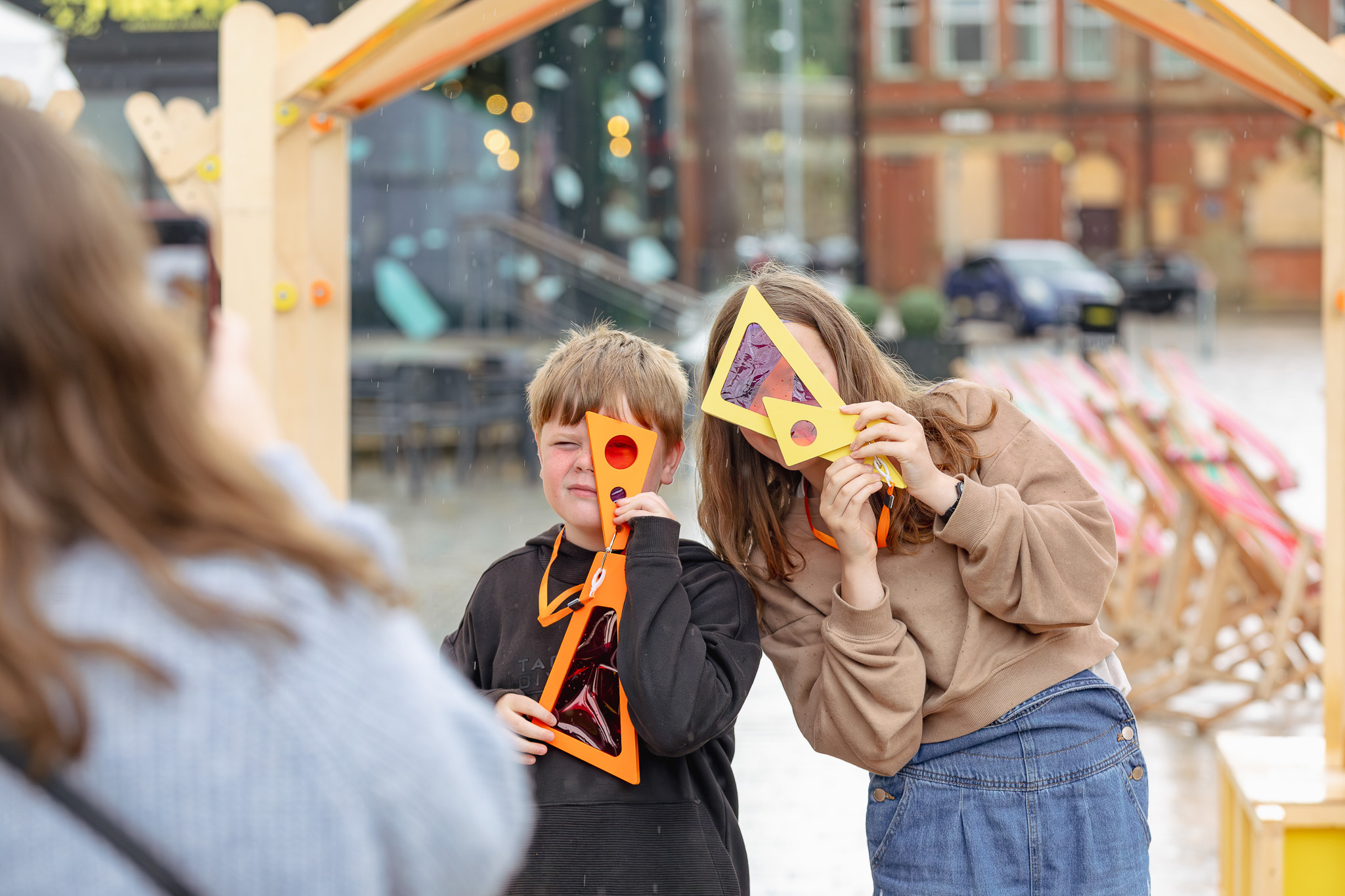 Children pose looking through colourful viewfinders held to their faces, in an outdoor setting.