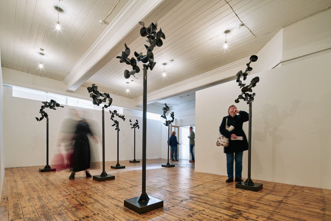 Gallery space with tall, abstract metal sculptures resembling trees. A blurred figure walks, and others observe.