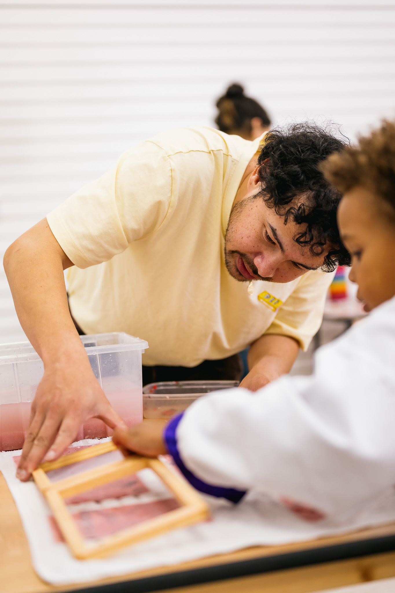 A man guides a child in a white coat as they work on a craft project using a wooden frame and water-filled container on a table.