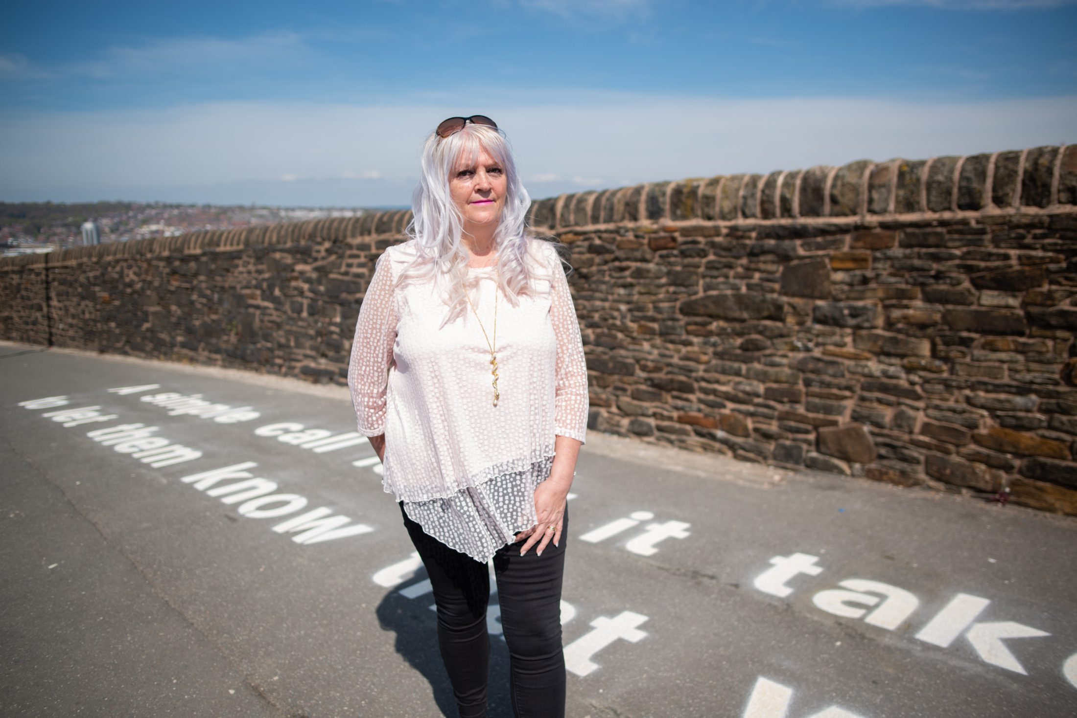 Woman with long hair stands on a sunny path by a stone wall. Text on the ground is partially visible.