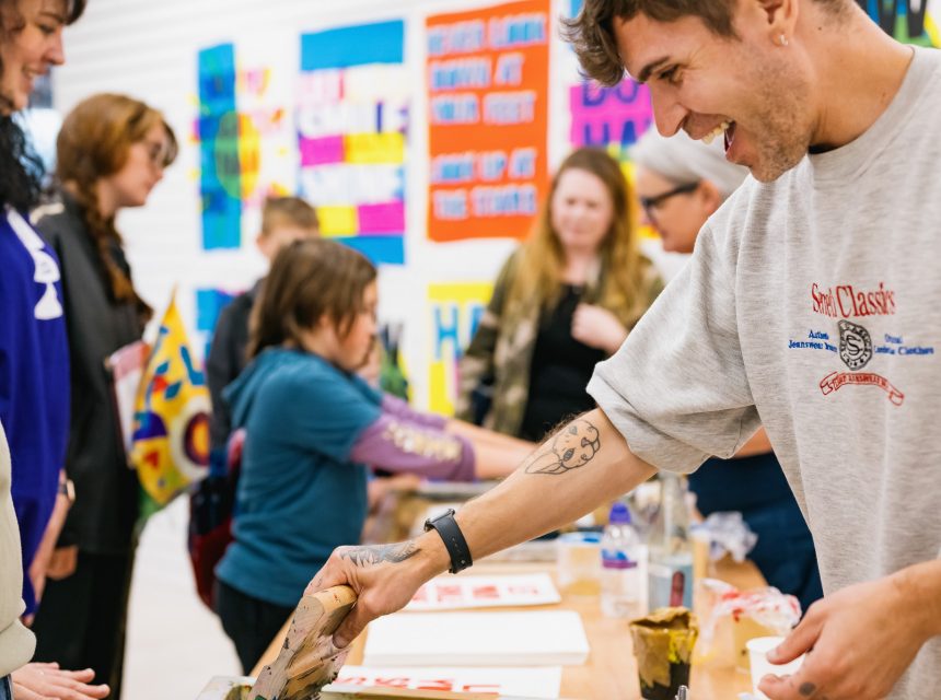 A smiling man prints artwork on paper whilst others, including adults and children, participate in an art workshop. Colourful posters decorate the background.