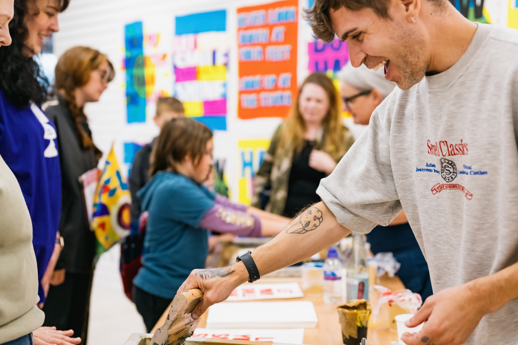 A smiling man prints artwork on paper whilst others, including adults and children, participate in an art workshop. Colourful posters decorate the background.