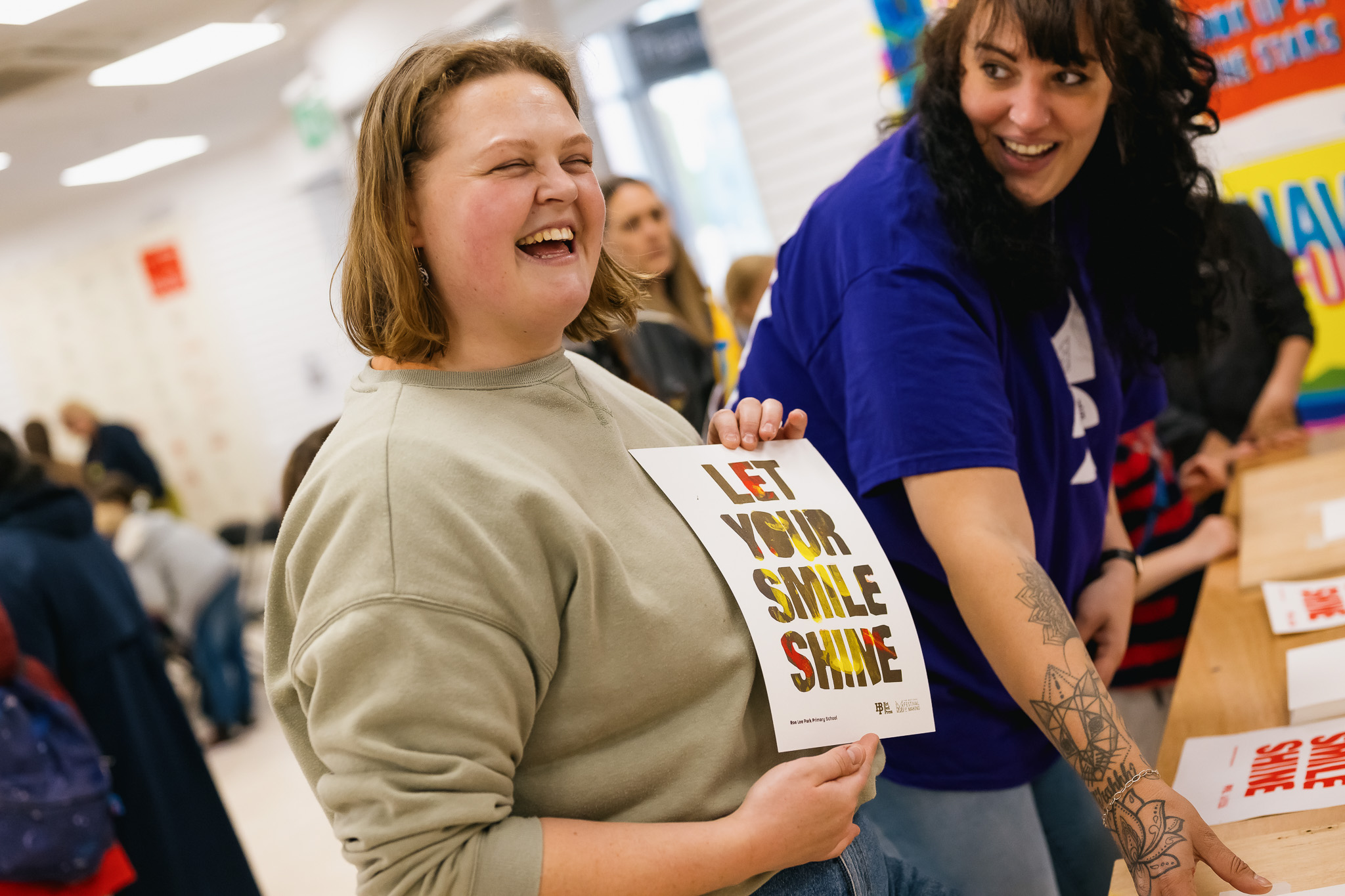 Two women smile at a table. One holds a sign that says LET YOUR SMILE SHINE. Other people are in the background, and the setting appears to be a cheerful, busy community event.