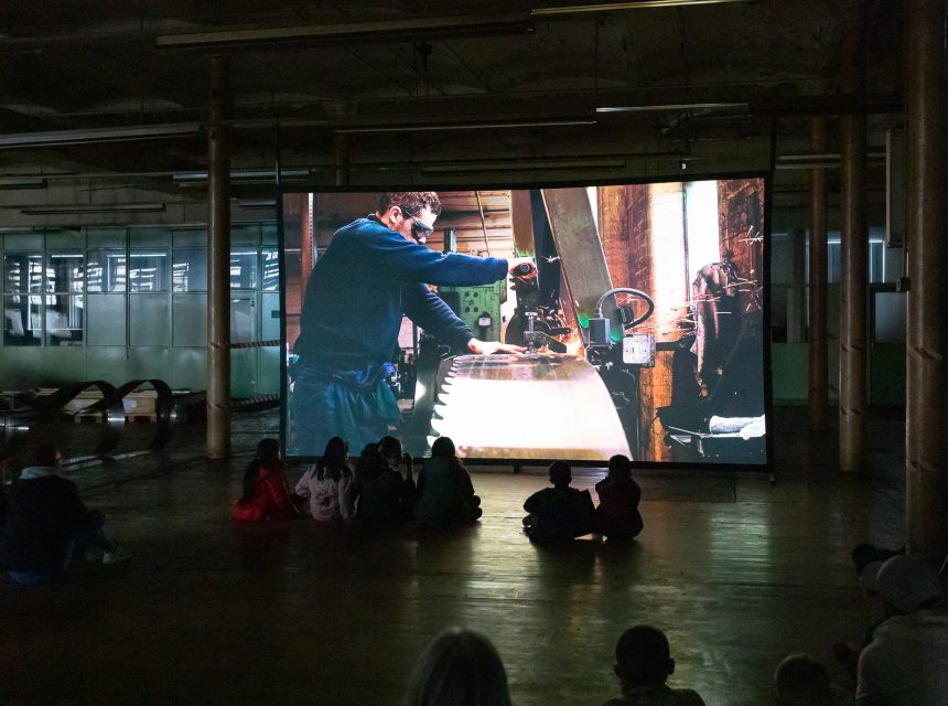A group of people sit on the floor in a dark room, watching a large screen that shows a video of a man working with a large tool or machine in a workshop.