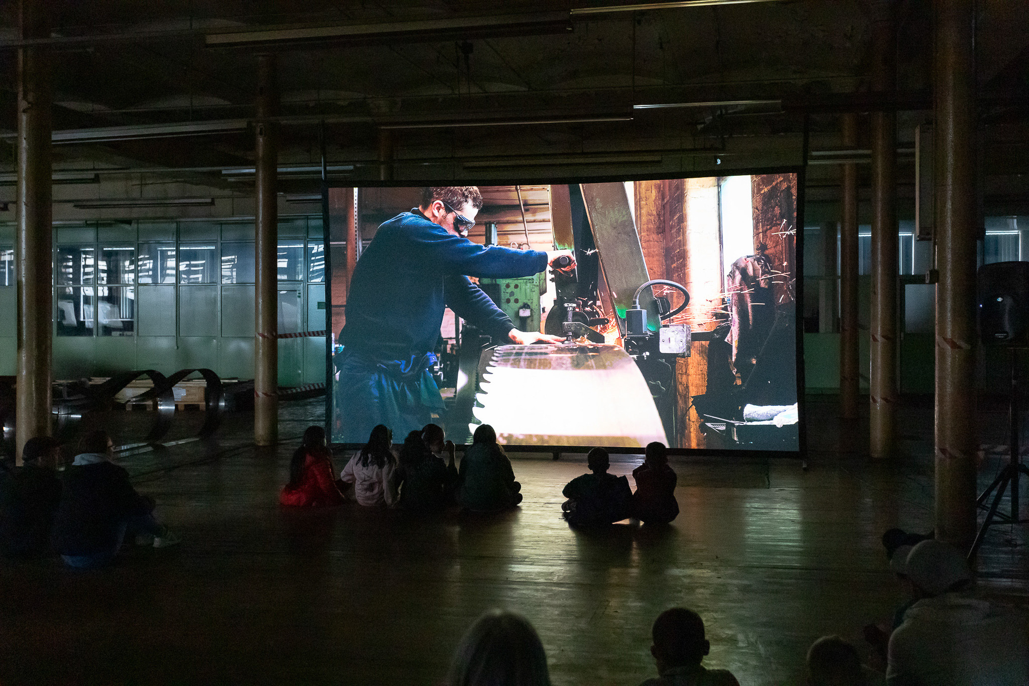 A group of people sit on the floor in a dark room, watching a large screen that shows a video of a man working with a large tool or machine in a workshop.