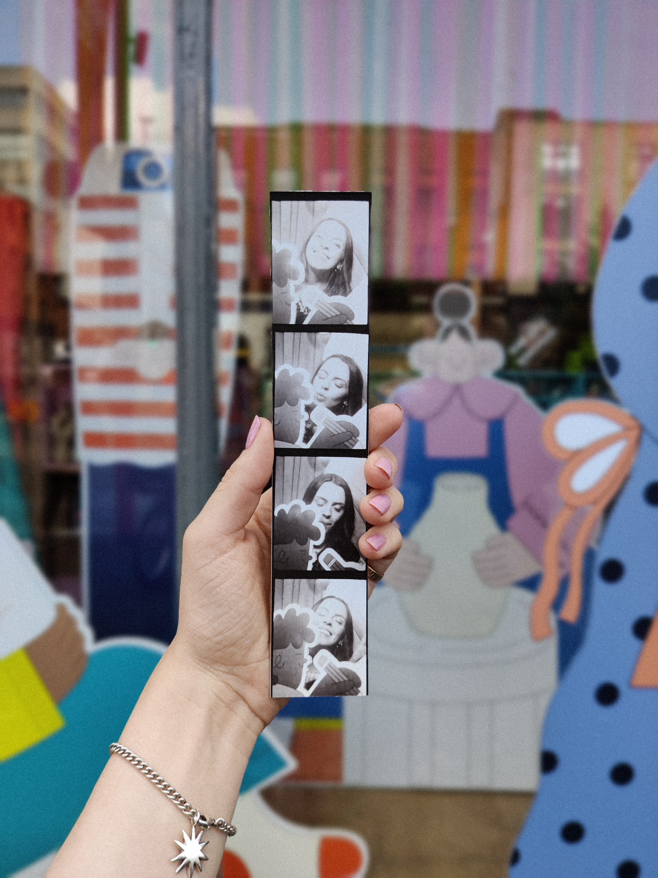 A hand with pink nails and a star bracelet holds a black-and-white photo strip showing a woman posing playfully. The background features colourful, cartoonish illustrations on a window.