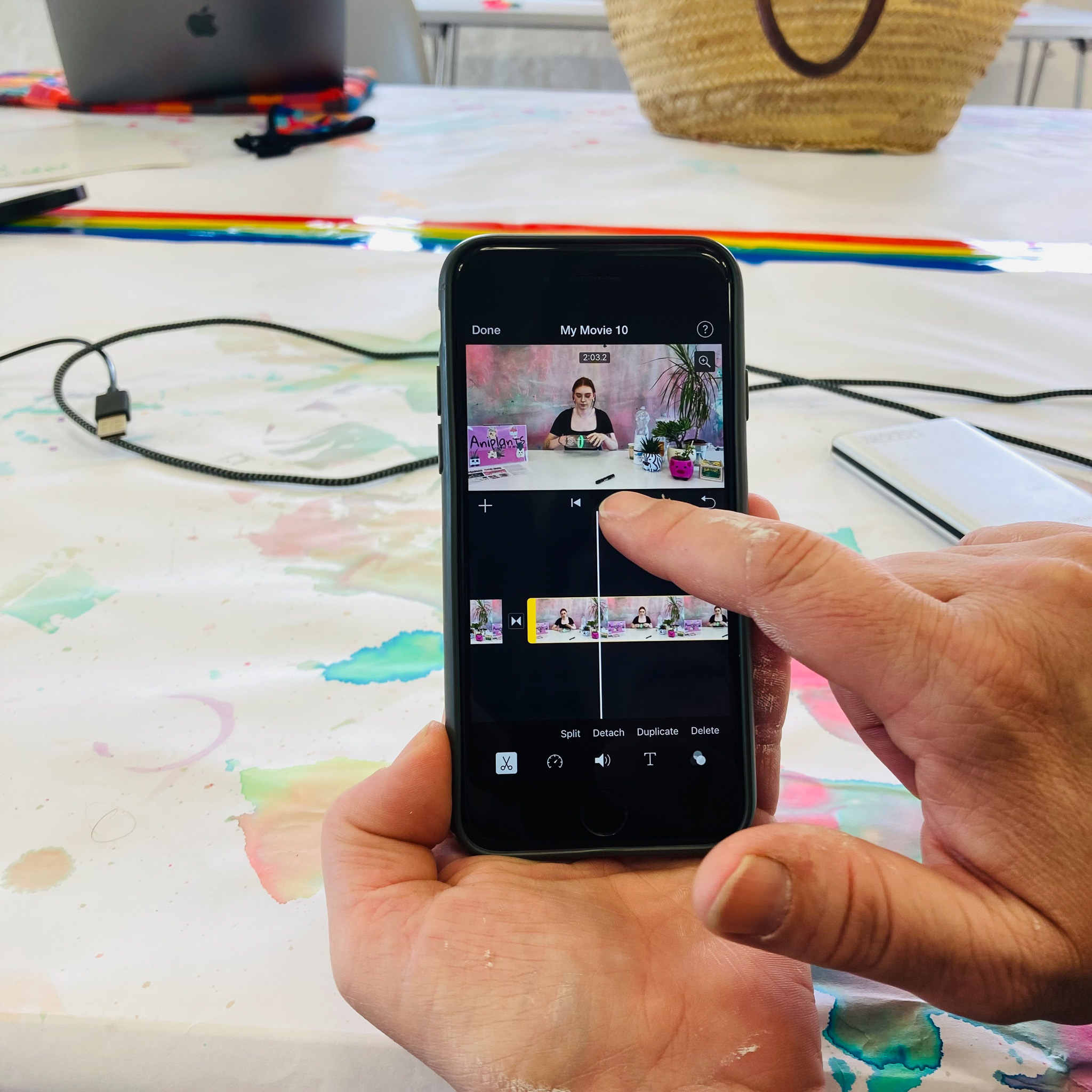 A person edits a video on a mobile using a video editing app. Their finger is touching the screen, and a messy table with art supplies and a laptop is visible in the background.