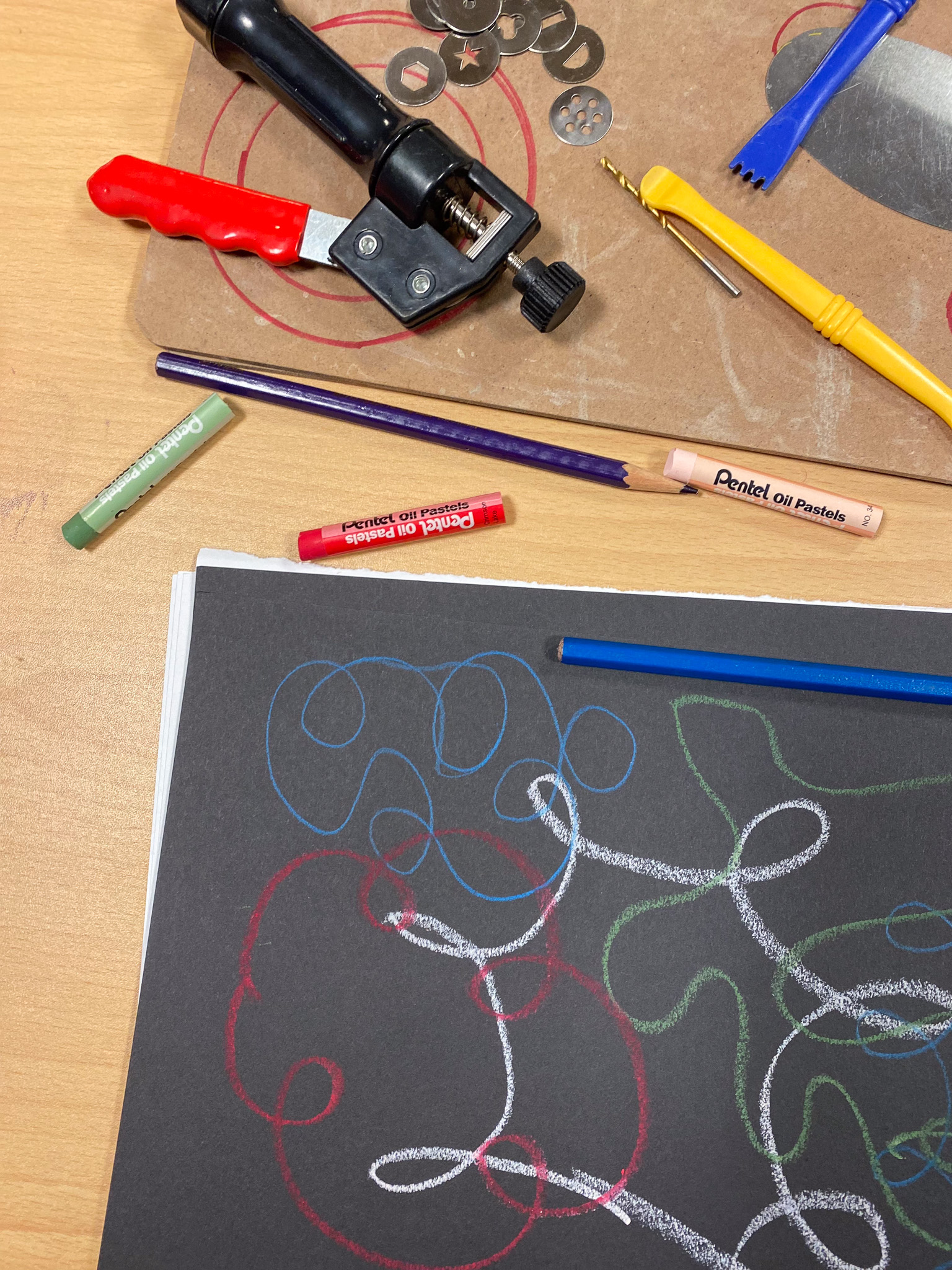 A creative workspace with colourful oil pastels and pencils; a black sheet shows abstract drawings, and clay tools rest on a cutting board.